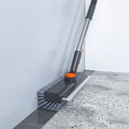 A Bangjie wall-mounted floor scrubber brush shown in use against a bathroom wall and floor, with foamy water visible. The product is dark gray with an orange accent and a black handle.
